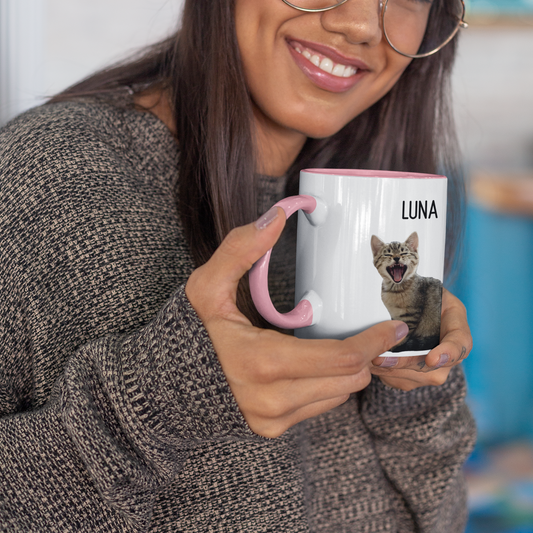 Smiling woman holding a personalized mug featuring a yawning tabby kitten and the name 'Luna'. The mug has a pink handle and interior, offering a custom gift option for pet lovers.