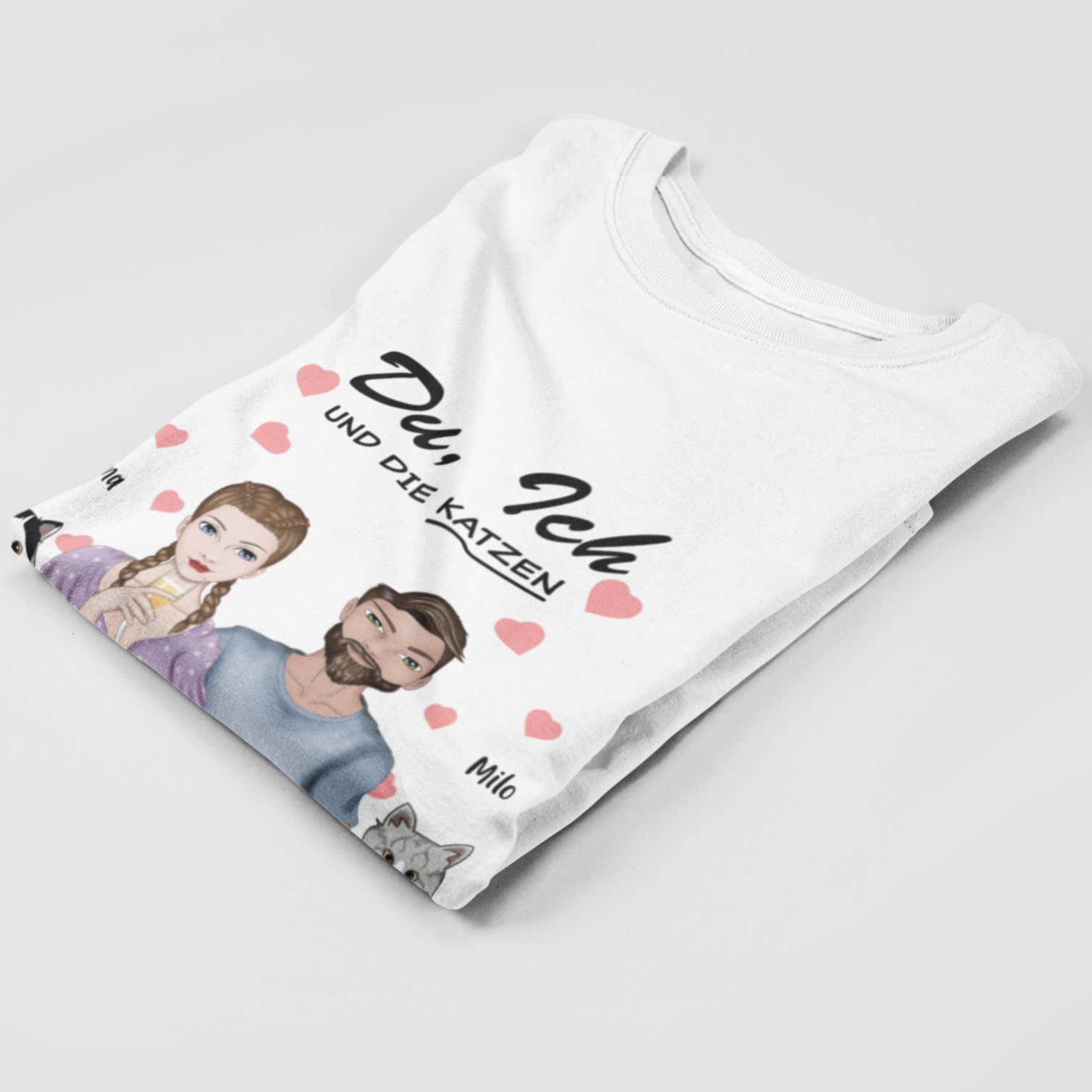 Folded t-shirt displaying the 'Du, ich und die Katzen' design with personalized woman and man characters and two custom cat images, each with their chosen name above.
